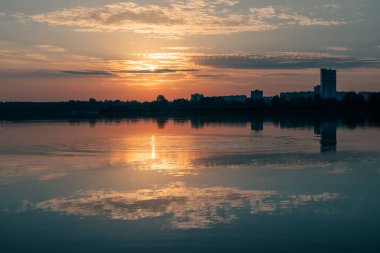 Bulutlu gökyüzünde, sakin göl suyuna yansıyan güneşli bir gün doğumu. Nehir kenarındaki şehir parkında huzurlu, mavi bir sabah