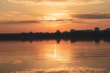 Bulutlu gökyüzünde, sakin göl suyuna yansıyan güneşli bir gün doğumu. Nehrin kenarındaki parkta altın bir sabah