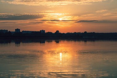 Bulutlu gökyüzünde, göl suyuna yansıyan güneşli bir gün doğumu. Şehir parkında nehir kenarında destansı bir sabah