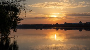 Bulutlu gökyüzünde, sakin göl suyuna yansıyan manzaralı sıcak gün doğumu. Nehir kenarındaki şehir parkında huzurlu bir sabah
