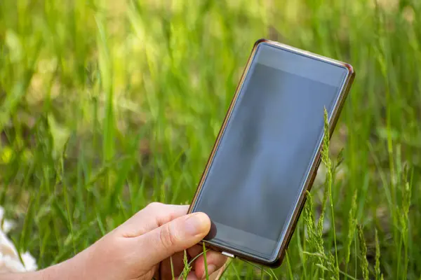 Holding smartphone with black screen in one hand in green field with blurry background, natural landscape mockup template