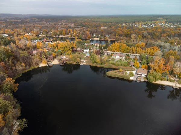 Aerial autumn cottages on river with colorful forest. Flying above vibrant autumnal village in Ukraine