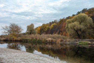 Kumlu nehir kıyısında sonbahar doğası bulutlu gökyüzü ve sakin su yüzeyinde ayna yansıması