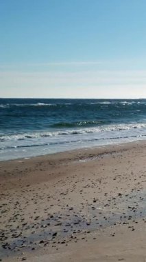 Güneşli deniz dalgaları boş bir kumsalda yuvarlanıyor, dikey deniz manzarası.