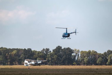 Blue Helicopter Robinson R44 Raven 1 flying above trees in blue sky on airshow above An-2 plane on the ground clipart