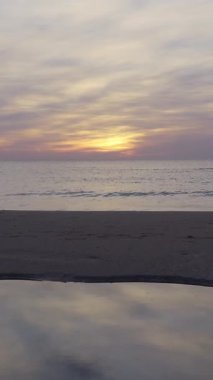 Deniz kıyısında gün doğumu manzarası, bulutlarda güneş ve kumların üzerinde sakin dalgalar.