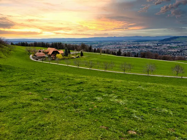 stock image Stunning sunset over a lush Swiss landscape, featuring a winding path through vibrant green fields leading towards the majestic snow-capped Alps, under a dramatic sky painted with hues of orange and blue