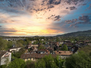 Gün batımında bir kasabanın panoramik manzarası. Köprüsü, çatıları, ağaçları ve tepeleri dramatik bir gökyüzü altında.