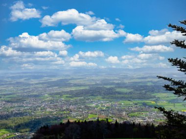 Bereketli bir vadinin panoramik manzarası kıvrımlı bir nehir, yeşil tarlalar, ve parçalı bulutlu mavi bir gökyüzü, bir yamaçtan görünen ağaçlarla ön planda