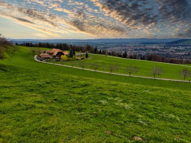 25.05.24.Switzerland. Gün batımında yeşil tarlaları, tepeleri ve dramatik bulutları olan bir şehrin panoramik manzarası