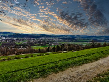 25.05.24.Switzerland. Gün batımında yeşil tarlaları, tepeleri ve dramatik bulutları olan bir şehrin panoramik manzarası