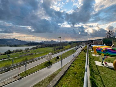 25.05.24.Switzerland. Bulutlu bir gökyüzünün altında, gölü ve dağları olan yeşil tarlaların arasından geçen bir yolun manzarası.