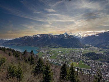 22.05.24 Switzerland. Geleneksel bir binası ve gün batımında karla kaplı dağları olan bir tepenin yanındaki göl kenti manzarası.