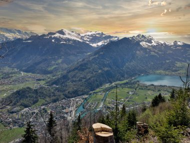 22.05.24 Switzerland. Geleneksel bir binası ve gün batımında karla kaplı dağları olan bir tepenin yanındaki göl kenti manzarası.