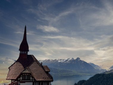 22.05.24 Switzerland. Geleneksel bir binası ve gün batımında karla kaplı dağları olan bir tepenin yanındaki göl kenti manzarası.