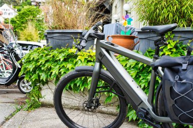 Arka planda geleneksel evler olan, yapraklı bir mahallede kaldırıma park etmiş, pannier çantalı modern elektrikli bisiklet.
