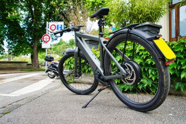 Arka planda geleneksel evler olan, yapraklı bir mahallede kaldırıma park etmiş, pannier çantalı modern elektrikli bisiklet.