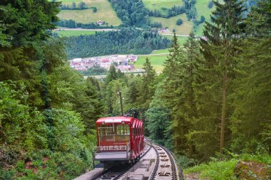 Kırmızı füniküler tren vagonu arka planda yeşil tarlalar ve köy manzaralı ormanlık bir alana iniyor.