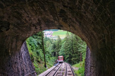 Kırmızı füniküler tren vagonu arka planda yeşil tarlalar ve köy manzaralı ormanlık bir alana iniyor.
