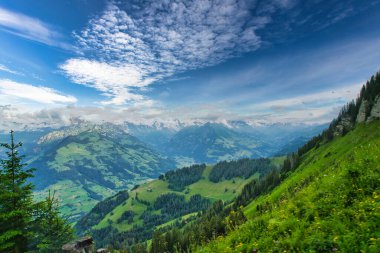 Parçalı bulutlu mavi gökyüzünün altında, arka planda karla kaplı tepeleri olan yeşil dağlar ve vadilerin görkemli manzarası.