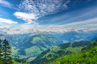 Parçalı bulutlu mavi gökyüzünün altında, arka planda karla kaplı tepeleri olan yeşil dağlar ve vadilerin görkemli manzarası.