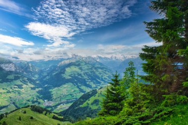 Parçalı bulutlu mavi gökyüzünün altında, arka planda karla kaplı tepeleri olan yeşil dağlar ve vadilerin görkemli manzarası.