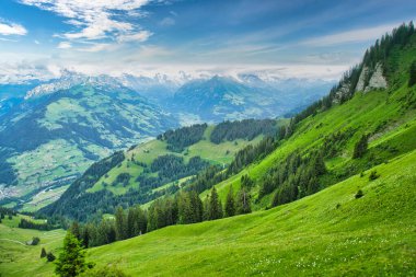 Parçalı bulutlu mavi gökyüzünün altında, arka planda karla kaplı tepeleri olan yeşil dağlar ve vadilerin görkemli manzarası.