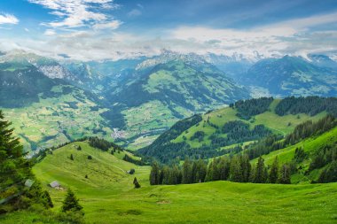 Parçalı bulutlu mavi gökyüzünün altında, arka planda karla kaplı tepeleri olan yeşil dağlar ve vadilerin görkemli manzarası.