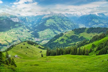 Parçalı bulutlu mavi gökyüzünün altında, arka planda karla kaplı tepeleri olan yeşil dağlar ve vadilerin görkemli manzarası.