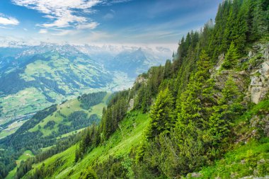Parçalı bulutlu mavi gökyüzünün altında, arka planda karla kaplı tepeleri olan yeşil dağlar ve vadilerin görkemli manzarası.