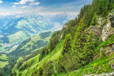 Parçalı bulutlu mavi gökyüzünün altında, arka planda karla kaplı tepeleri olan yeşil dağlar ve vadilerin görkemli manzarası.