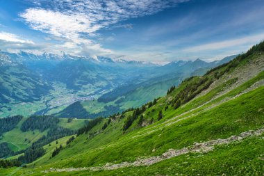 Parçalı bulutlu mavi gökyüzünün altında, arka planda karla kaplı tepeleri olan yeşil dağlar ve vadilerin görkemli manzarası.