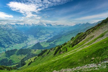Parçalı bulutlu mavi gökyüzünün altında, arka planda karla kaplı tepeleri olan yeşil dağlar ve vadilerin görkemli manzarası.