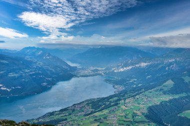 Dağlarla ve vadilerle çevrili, parçalı bulutlu mavi gökyüzünün altında dağınık köylerle panoramik bir göl manzarası.