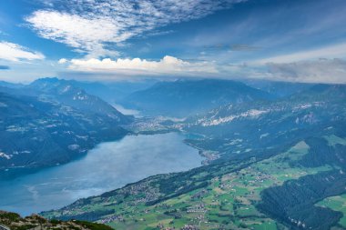 Dağlarla ve vadilerle çevrili, parçalı bulutlu mavi gökyüzünün altında dağınık köylerle panoramik bir göl manzarası.