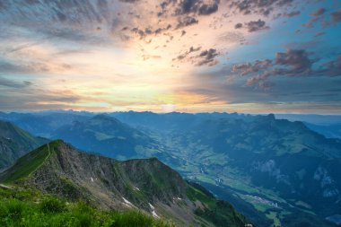 Gün batımında, dramatik bulutlar ve vadilerle çevrili, İsviçre bayrağı ve dağ kulübesinde nefes kesici bir dağ zirvesi.