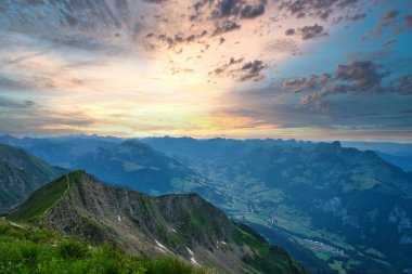 Gün batımında, dramatik bulutlar ve vadilerle çevrili, İsviçre bayrağı ve dağ kulübesinde nefes kesici bir dağ zirvesi.