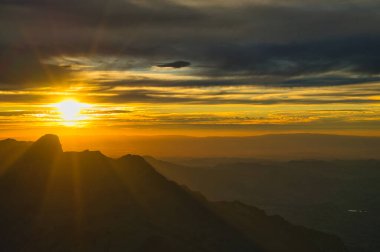 Güneş ışınlarının bulutları delip geçtiği ve manzarayı aydınlattığı sıradağların üzerinden gün batımının çarpıcı görüntüsü.