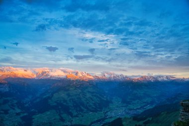 Güneş doğarken karla kaplı dağların panoramik manzarası, altın ışıkla yıkanmış tepeler ve yukarıda dramatik bir gökyüzü