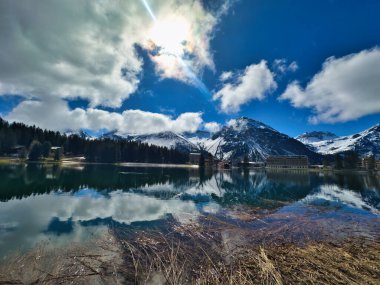Dağlarla çevrili sakin göl karla kaplı tepeler ve sudaki manzaranın yansımaları. Sahne bulutların arasından yükselen parlak bir güneş ile güçlenir..