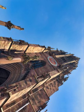 Karmaşık detayları olan gotik bir mimari ve açık mavi gökyüzünün altında belirgin bir kule. Güneş manzaraya parlak bir ışık katıyor, binaların canlı renklerini güçlendiriyor.