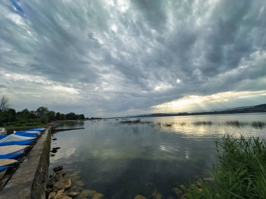 Dramatik ve bulutlu bir gökyüzü altında sakin bir göl manzarası. Güneş ışınları bulutları delip geçiyor, sakin sulara ışık tutuyor, kıyıya yanaşmış kayıklar ve yemyeşil su kenarlarını kaplıyor.