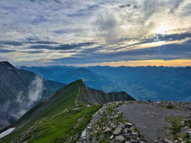Alacakaranlıkta muhteşem bir dağ manzarası. Kayalık tepeye çıkan dolambaçlı bir patika. Gökyüzü mavi ve turuncu renklerle boyanıyor güneş uzak tepelerin ardında batarken nefes kesici bir manzara yaratıyor.