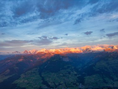 Alacakaranlıktaki bir dağ sırasının çarpıcı manzarası. Tepeler batan güneşten gelen sıcak, altın bir ışıkla yıkanmış. Gökyüzü mavi ve turuncu renklerin bir karışımıdır, aşağıdaki engebeli araziye karşı güzel bir kontrastlık yaratır.