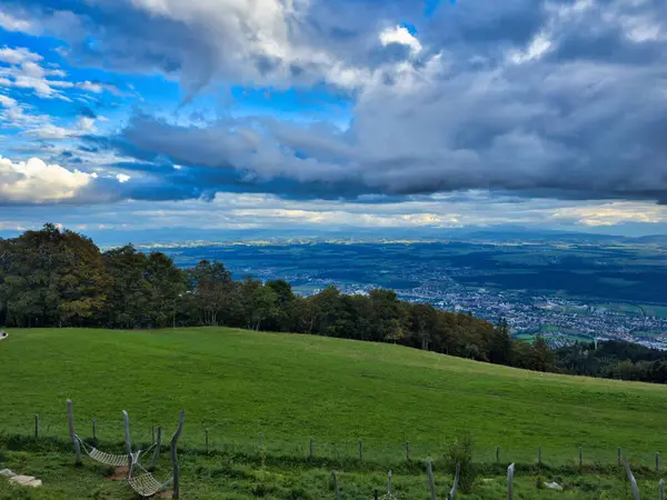 stock image A picturesque rural landscape featuring green fields under a partly cloudy sky, with a farmstead and a modern barn in the background, surrounded by gentle rolling hills and forested areas