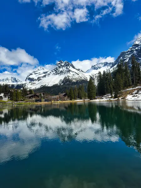 stock image Serene lake surrounded by mountains with snow-capped peaks and reflections of the landscape in the water. The scene is enhanced by a bright sun breaking through the clouds.