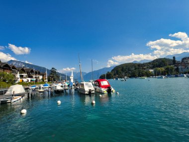 28.07.24 Switzerland. Berrak mavi gökyüzünün altında dağlarla ve evlerle çevrili sakin mavi bir göle yanaşmış tekneli bir marina manzarası.