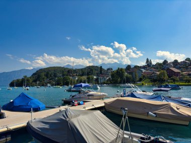 28.07.24 Switzerland. Berrak mavi gökyüzünün altında dağlarla ve evlerle çevrili sakin mavi bir göle yanaşmış tekneli bir marina manzarası.