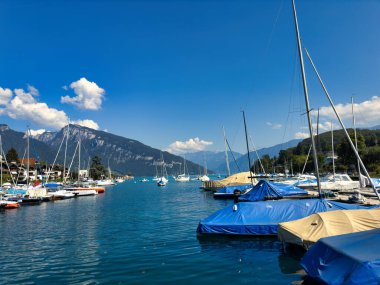 28.07.24 Switzerland. Berrak mavi gökyüzünün altında dağlarla ve evlerle çevrili sakin mavi bir göle yanaşmış tekneli bir marina manzarası.