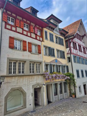 28.07.24 Switzerland. Tarihi binaları ve renkli mimarisi olan bir Avrupa şehrinde rahat bir cadde. Sahnede ön planda dekoratif bir çeşme ve eski evlerin büyüleyici cepheleri yer alıyor..